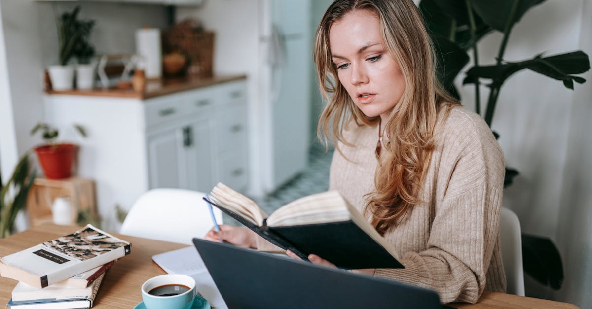 Young focused female studying planner near netbook and cup of hot aromatic coffee on blurred background of kitchen interior