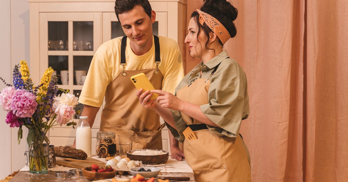 A couple in aprons preparing food and using a smartphone in a cozy kitchen setting.