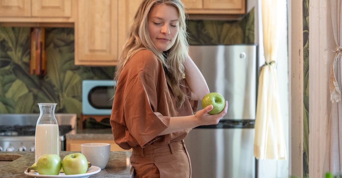 A caucasian woman enjoys a moment in an indoor kitchen while holding a green apple and surrounded by fresh decor.