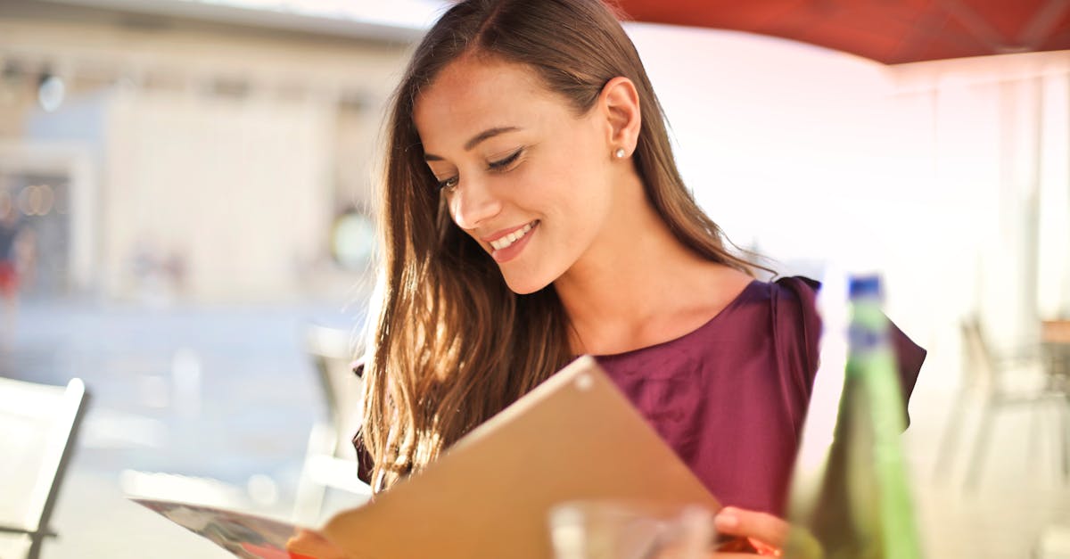 A woman smiles as she reads a menu at an outdoor restaurant, enjoying a sunny day.