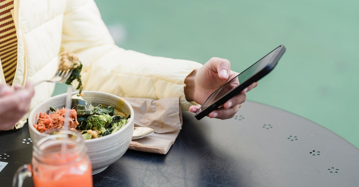 Crop unrecognizable female with cellphone sitting at cafe table with delicious vegan salad and smoothie in daylight